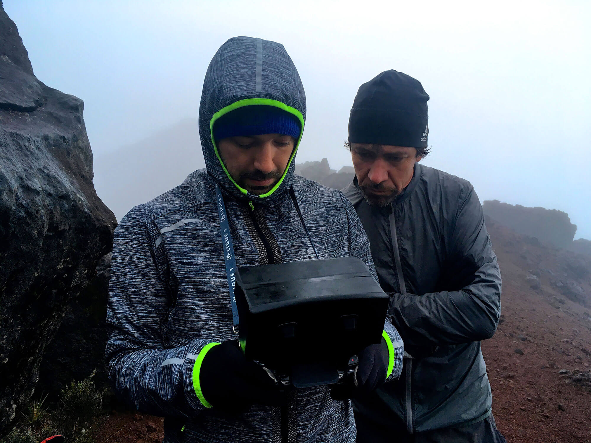 Photographie d'Émile Rouet, pilote de drone, et de Laurent Bouit, réalisateur de la série de documentaires Les Plus Beaux Treks, entrain de regarder l'écran de contrôle du drone, en haut du Python des Neiges, sur l'Île de la Réunion. 